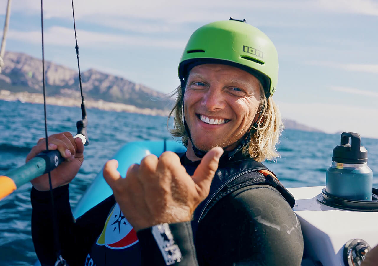 Jannis Maus, der einen grünen Helm und einen Neoprenanzug trägt, lächelt und macht ein Shaka-Zeichen, während er auf dem Meer segelt, während im Hintergrund die Berge und die Küstenlinie zu sehen sind.