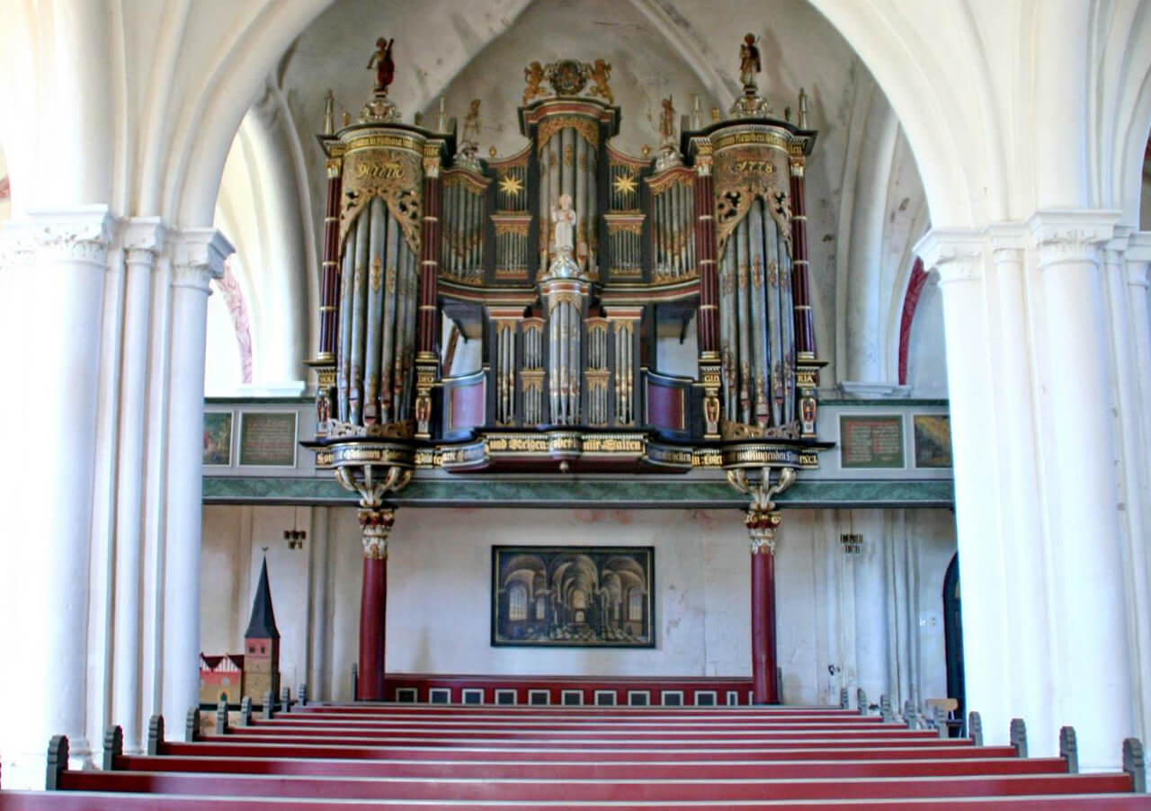 Eine große, verzierte Pfeifenorgel mit goldenen und roten Akzenten ist über den Kirchenbänken in einer Kirche mit gewölbten weißen Säulen und gewölbten Decken angebracht. Unter der Orgel sind Kunstwerke und ein Modellbau zu sehen.
