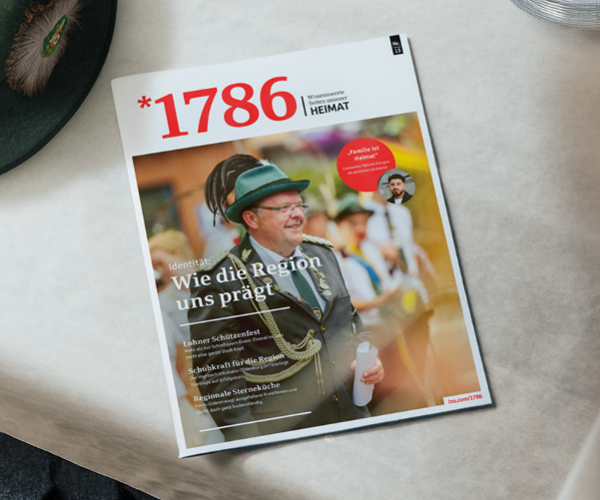 Das Magazin mit dem Titel 1786, auf deren Titelseite ein Mann in traditioneller Schützenfestuniform abgebildet ist, liegt auf einem weißen Tisch neben einem Glas Wasser und einem grünen Schützenfest-Hut mit Feder.