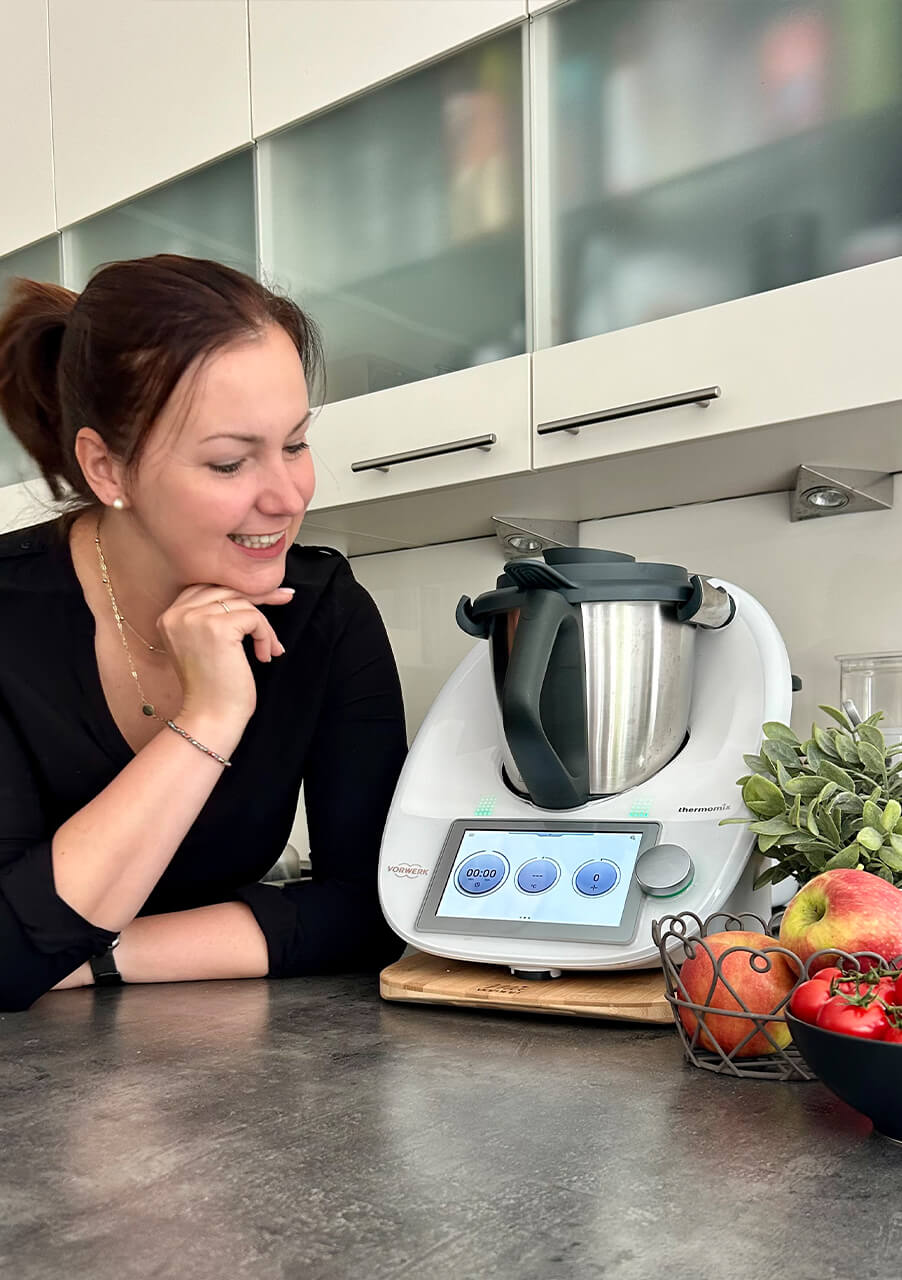 Eine Frau in einem schwarzen Oberteil lächelt, während sie ein Küchengerät mit einem digitalen Bildschirm auf einer Theke betrachtet, umgeben von frischem Obst, Gemüse und einer kleinen Pflanze.