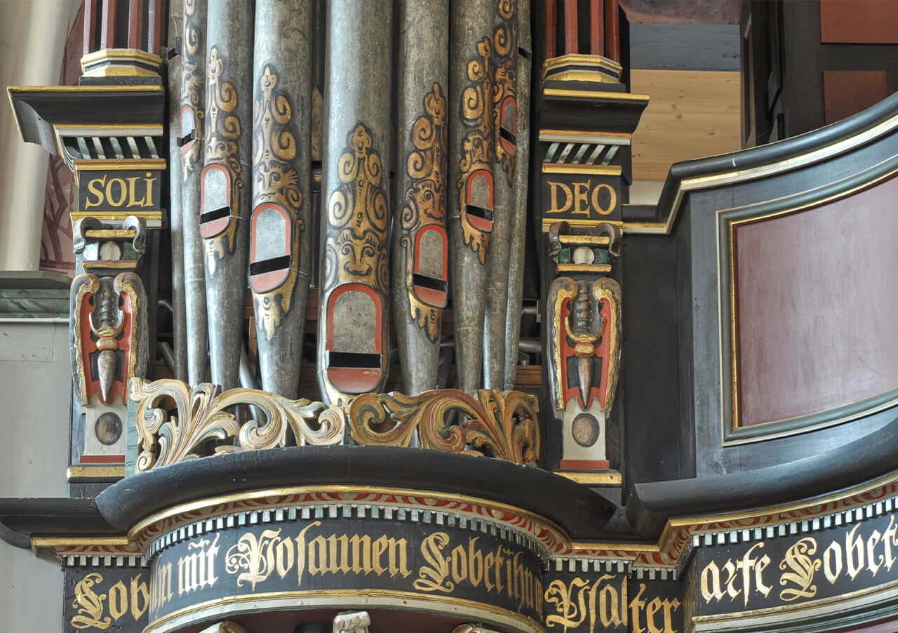 Nahaufnahme von verschnörkelten Kirchenorgelpfeifen mit dekorativen Details in Gold, Rot und Schwarz. Auf dem geschwungenen Holzrahmen der Orgel sind die Worte Soli Deo und andere deutsche Sprüche eingraviert.