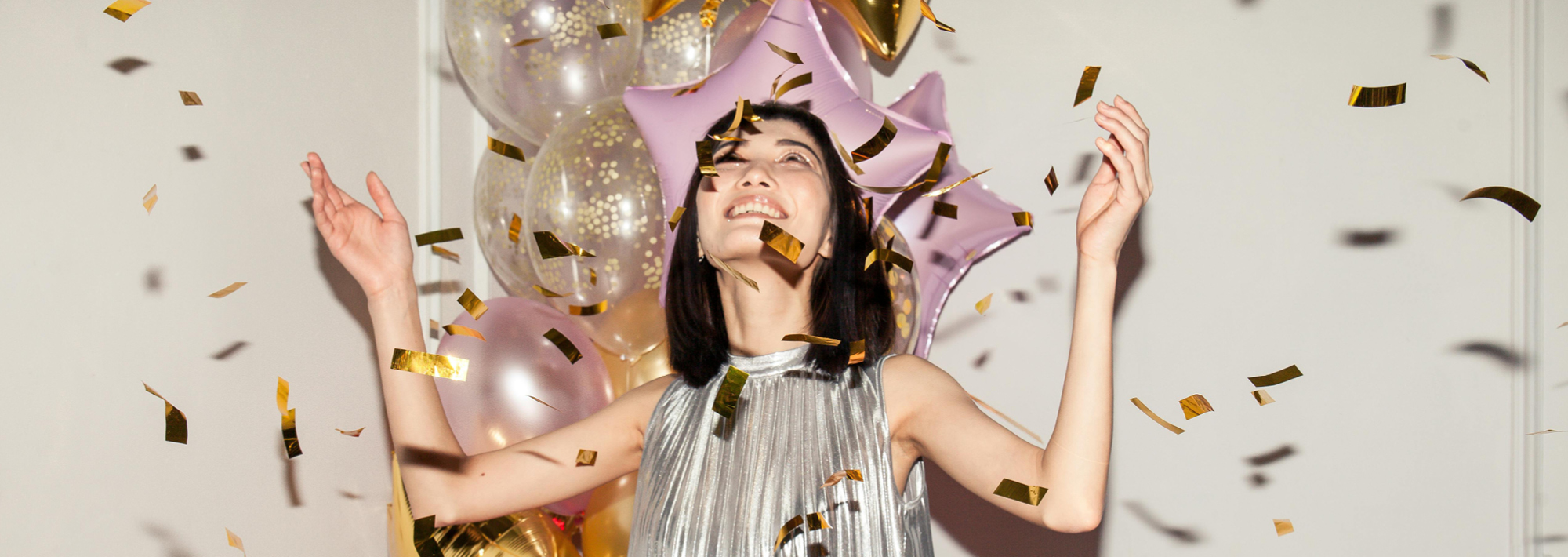 Eine Frau in einem silbernen Kleid lächelt und reißt die Arme hoch, während um sie herum goldenes Konfetti fällt. Hinter ihr befinden sich rosafarbene sternförmige Luftballons und durchsichtige Luftballons, die eine festliche Atmosphäre schaffen.