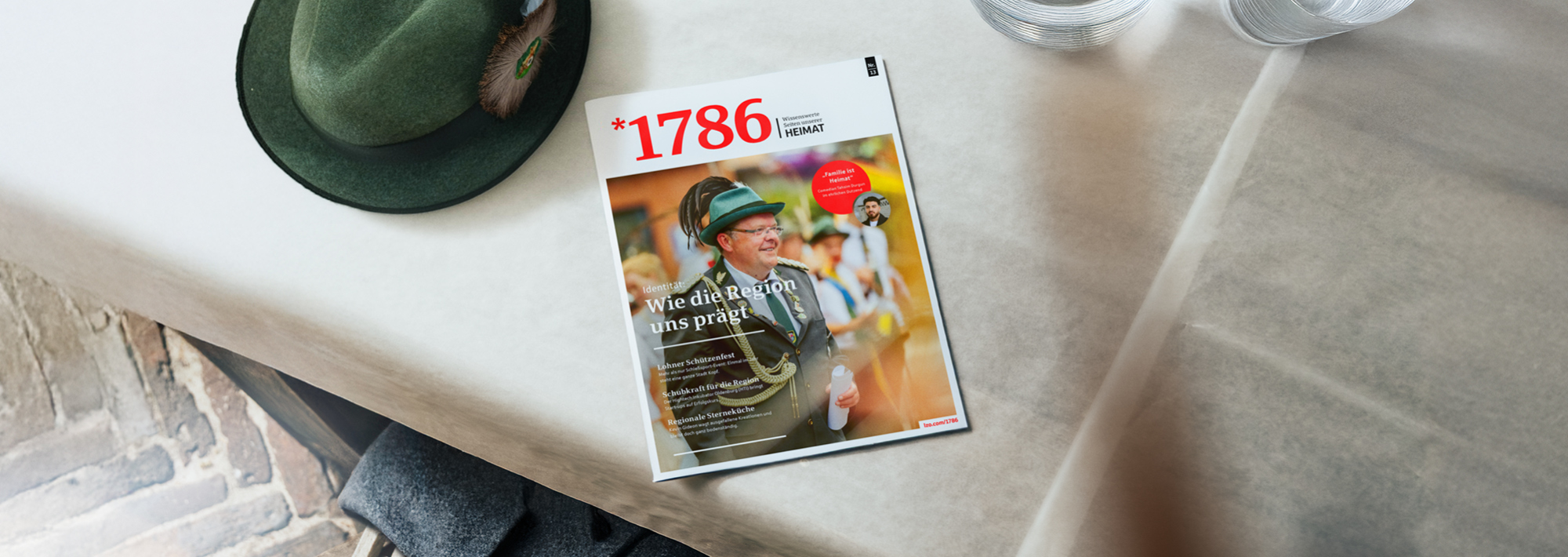 Das Magazin mit dem Titel 1786, auf deren Titelseite ein Mann in traditioneller Schützenfestuniform abgebildet ist, liegt auf einem weißen Tisch neben einem Glas Wasser und einem grünen Schützenfest-Hut mit Feder.