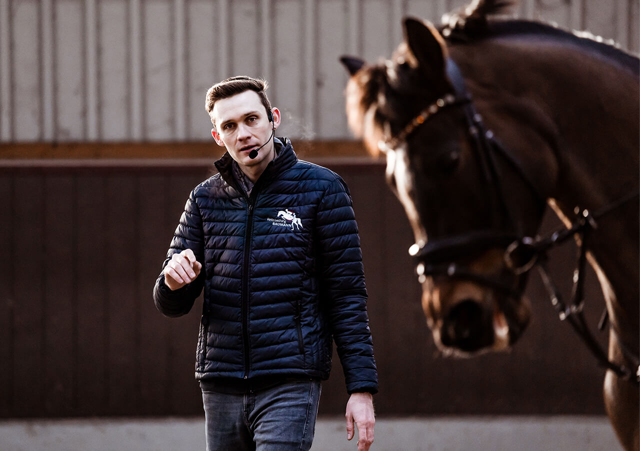 Ein Mann mit Headset-Mikrofon und Steppjacke steht in einem Innenraum und gestikuliert mit der linken Hand, während er über mentale Stärke spricht. In der Nähe steht ein braunes Pferd im Zaumzeug leicht unscharf.