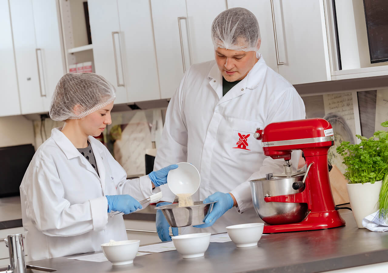 Zwei Personen in Laborkitteln, Haarnetzen und Handschuhen arbeiten in einem Küchenlabor und demonstrieren Produktinnovation und Transformation, indem sie Zutaten in eine Schüssel schütten, die von anderen weißen Schüsseln, einem roten Standmixer und einer nahe gelegenen Pflanze umgeben ist.