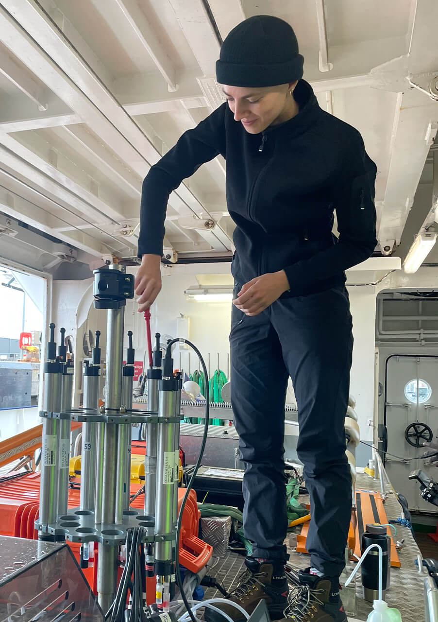 Eine Person in schwarzer Outdoor-Kleidung steht auf einer Bank im Inneren eines Schiffes und benutzt ein Werkzeug, um wissenschaftliche Geräte mit Metallzylindern einzustellen - vielleicht denkt sie über Karrierewechsel und Sinnfindung nach, während sie sich auf ein Forschungsexperiment vorbereitet.