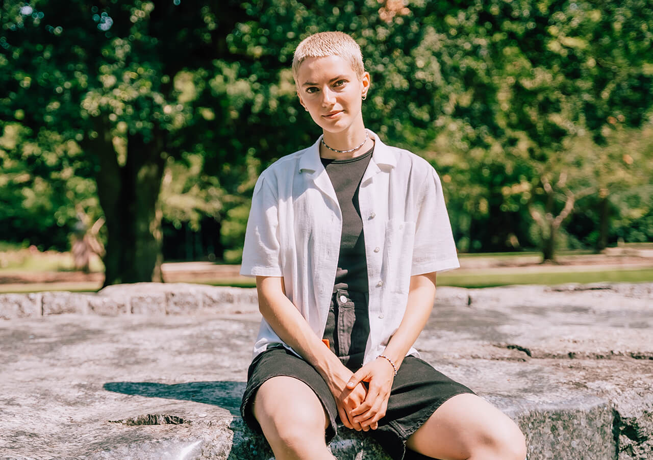 Eine Person mit kurzen blonden Haaren sitzt auf einer Steinfläche im Freien und denkt über Karrierewechsel und Sinnfindung nach. Sie trägt ein weißes kurzärmeliges Hemd über einem schwarzen T-Shirt und Shorts. Grüne Bäume und Sonnenlicht erhellen den friedlichen Hintergrund.