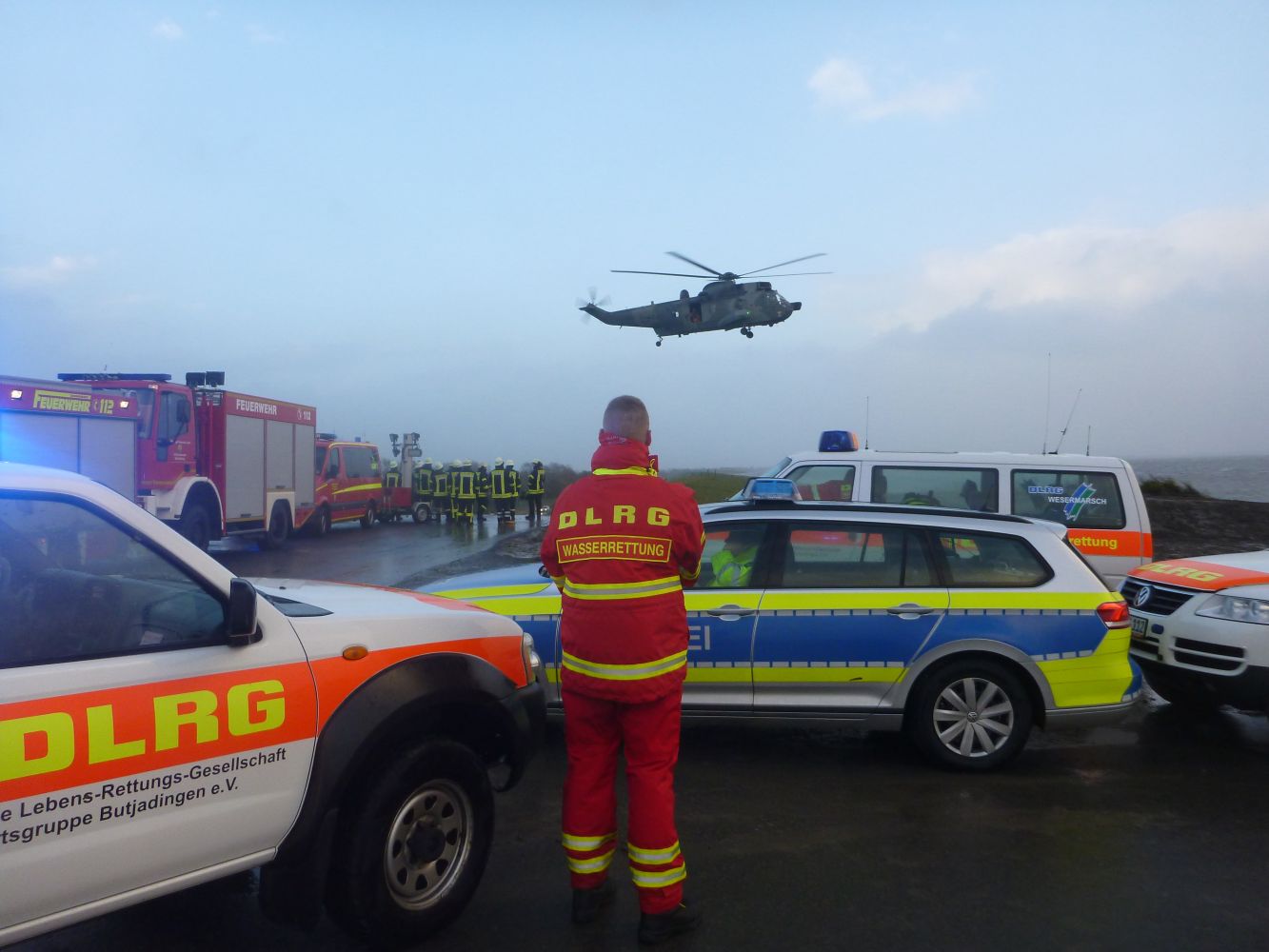 Ein Rettungssanitäter in einer rot-gelben DLRG-Uniform steht inmitten von Einsatzfahrzeugen, während ein Hubschrauber über dem Schauplatz in Küstennähe schwebt, während im Hintergrund weitere Einsatzkräfte und Feuerwehrfahrzeuge zu sehen sind.