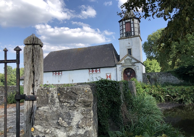 Eine kleine weiße Kirche mit einem Holzdach und einem hohen Glockenturm steht hinter einer Steinmauer und einem Tor, umgeben von Grün unter einem teilweise bewölkten blauen Himmel.