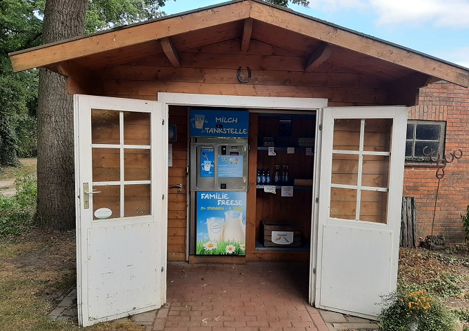 Ein kleiner Holzschuppen mit offenen Flügeltüren beherbergt einen Automaten mit der Aufschrift "Milch Tankstelle" für frische Milch sowie verschiedene Milchprodukte, die in Regalen ausgestellt sind. Der Schuppen steht an einem gemauerten Weg in einer ländlichen Umgebung.