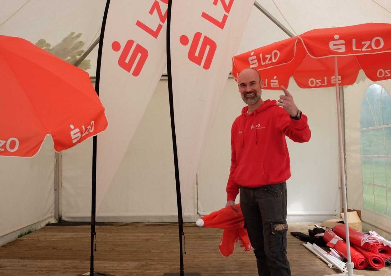 Ein Mann in einem roten Kapuzenpulli steht lächelnd unter zwei rot-weißen Regenschirmen und Bannern mit dem LzO-Logo in einem Zelt und hält ein Handy für ein Selfie in die Hand. In der Nähe liegen Stoffrollen auf dem Boden.