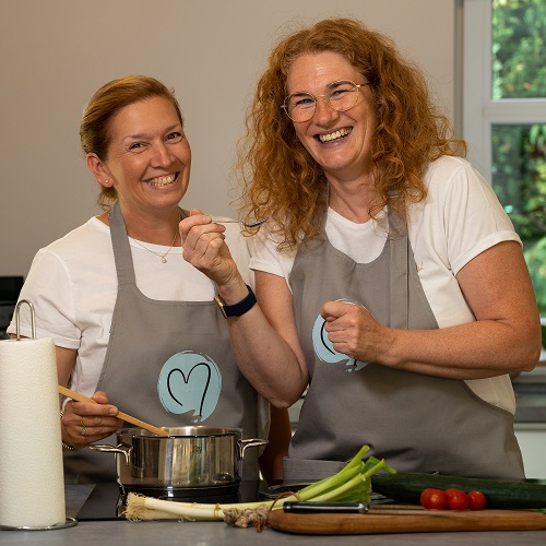 Zwei Frauen in grauen Schürzen lächeln, während sie gemeinsam in einer Küche kochen. Die eine rührt in einem Topf, die andere hält einen Löffel. Frisches Gemüse und eine Papierhandtuchrolle liegen vor ihnen auf der Theke.