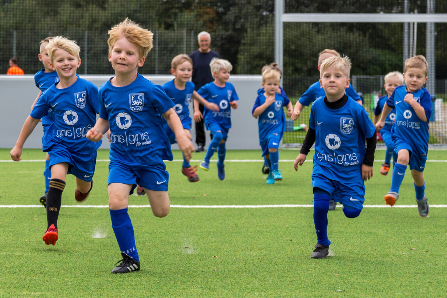 Eine Gruppe von Jungen in blauen Fußballtrikots rennt energisch über einen grünen Fußballplatz, lächelt und hat Spaß. Im Hintergrund sind ein Trainer und Torpfosten zu sehen - ein Mehrzweckplatz für alle Generationen.