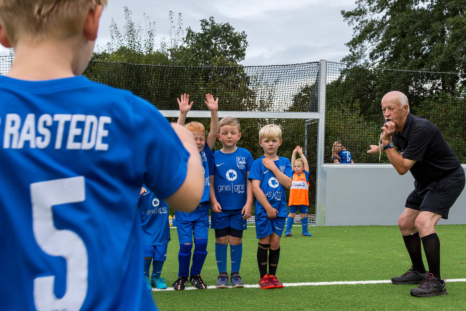 Eine Gruppe junger Kinder in blauen Fußballtrikots steht in einer Reihe auf einem Fußballfeld, während ein schwarz gekleideter Trainer Anweisungen gibt, was Mehr Platz für alle Generationen illustriert. Ein Kind im Vordergrund trägt ein Trikot mit der Aufschrift "RASTEDE 5" auf dem Rücken.