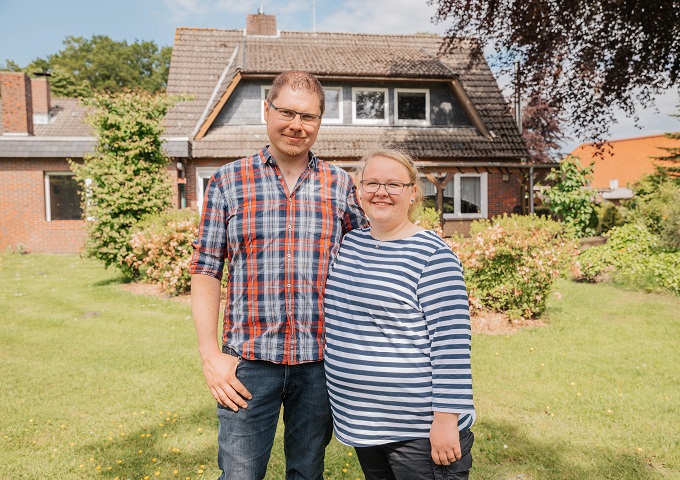 Ein Mann und eine Frau stehen an einem sonnigen Tag lächelnd vor ihrem renovierten Backsteinhaus nach einer Sanierung Eigenheim in Westerstede, mit Garten und grünem Rasen. Der Mann trägt ein kariertes Hemd, die Frau trägt ein gestreiftes Hemd und eine Brille.