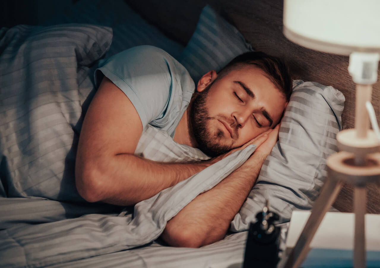 Ein junger Mann mit Bart liegt auf der Seite im Bett und stützt seinen Kopf auf seine Hand. Er ist mit einer gestreiften Decke zugedeckt, und neben ihm ist eine Nachttischlampe eingeschaltet.