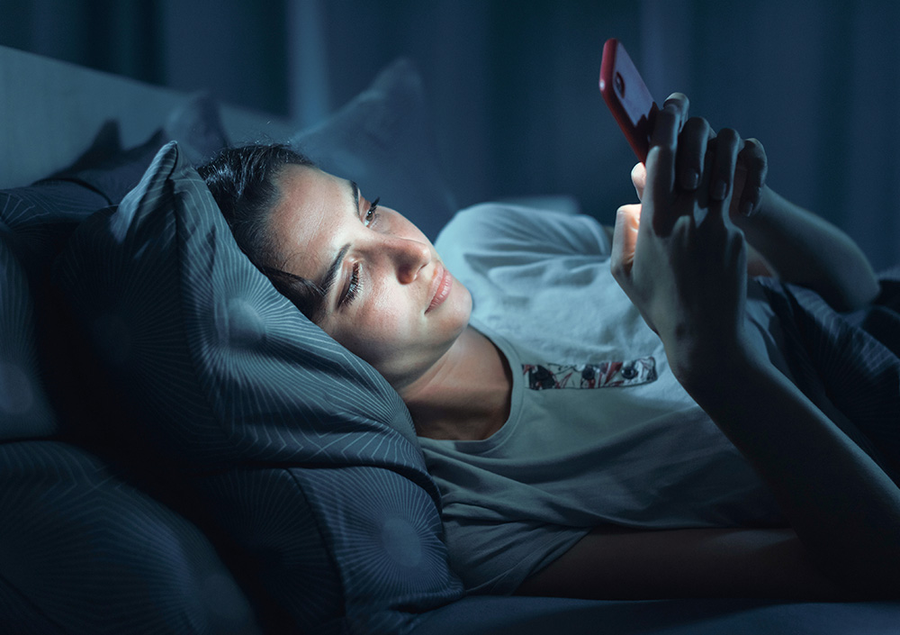 Eine Frau liegt nachts im Bett und schaut auf ihr Smartphone, das von seinem Bildschirm beleuchtet wird, während ihr Kopf auf einem Kissen ruht und das Zimmer nur schwach beleuchtet ist.