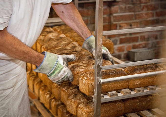 Eine Person mit Handschuhen und weißer Kleidung ordnet in einer Bäckerei mit Sichtbacksteinwänden frische Brote auf Metallgestellen an.