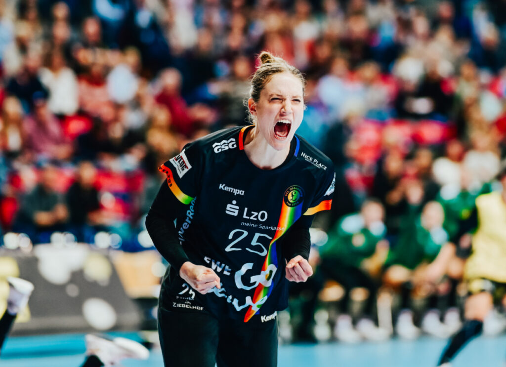 Torhüterin Madita Kohorst, die ein schwarzes Trikot mit der Nummer 25 trägt, schreit leidenschaftlich und ballt während eines Spiels die Fäuste. Im Hintergrund sind eine unscharfe Menge und andere Spielerinnen zu sehen.
