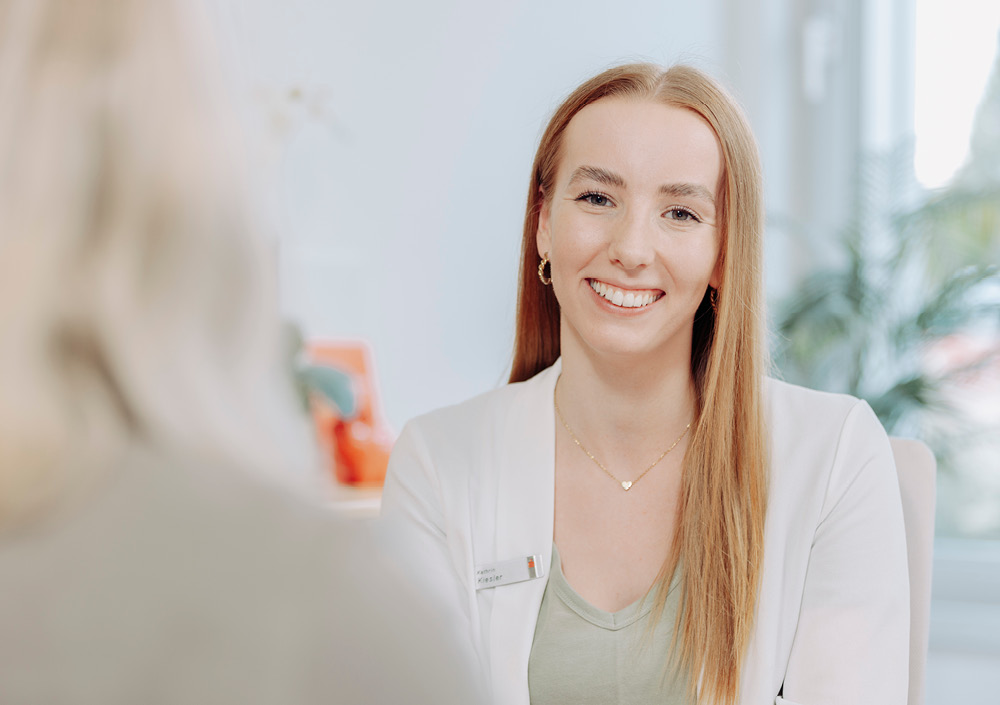 Eine Frau mit langem, glattem Haar, weißem Blazer und Namensschild lächelt, während sie einer anderen Person in einem Büro mit Tageslicht und Pflanzen im Hintergrund gegenübersitzt.