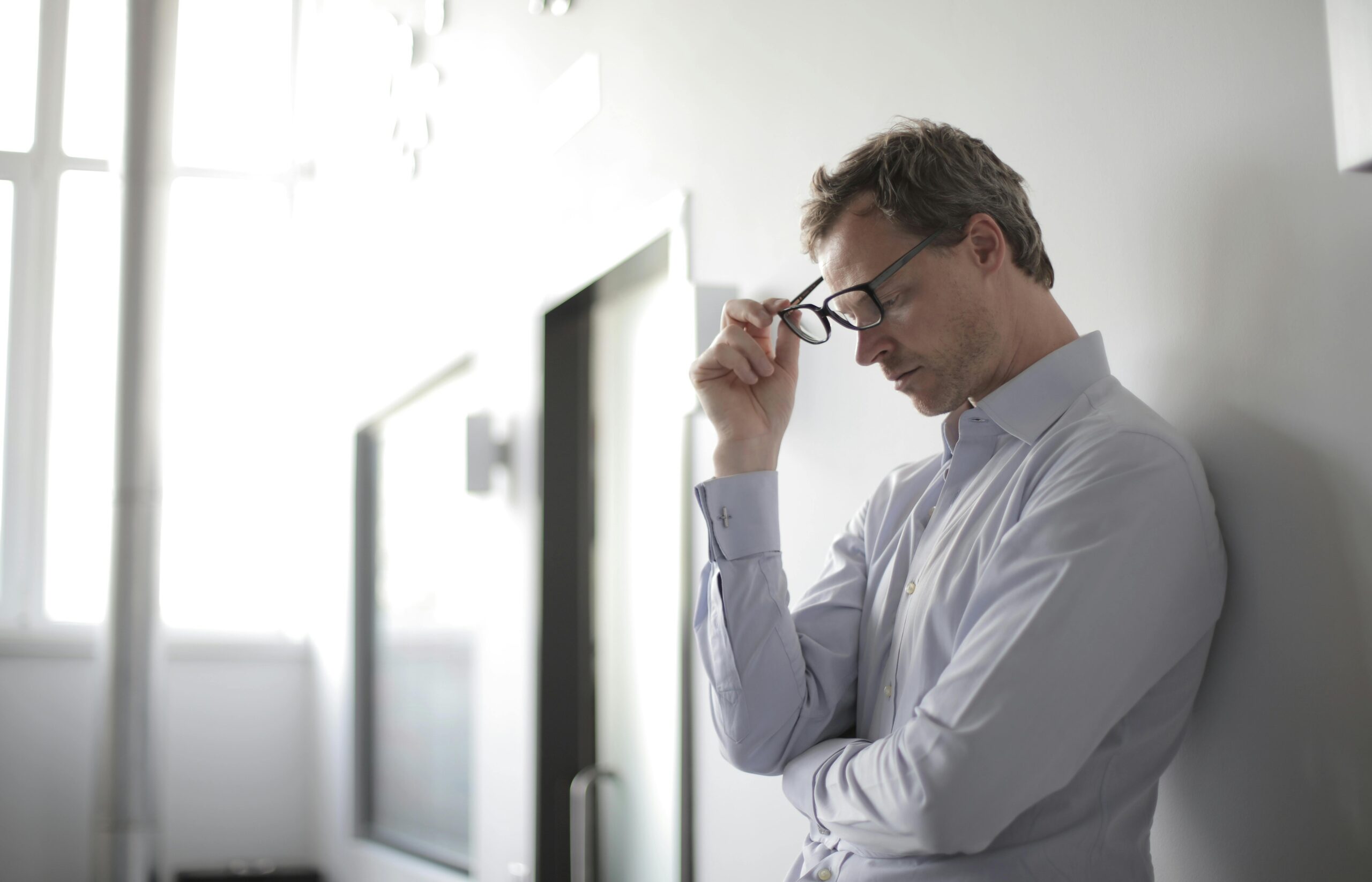 Ein Mann in einem hellblauen Hemd steht in einem Haus, lehnt mit geschlossenen Augen und einer Brille in der Hand an einer Wand und scheint in Gedanken versunken oder gestresst zu sein.