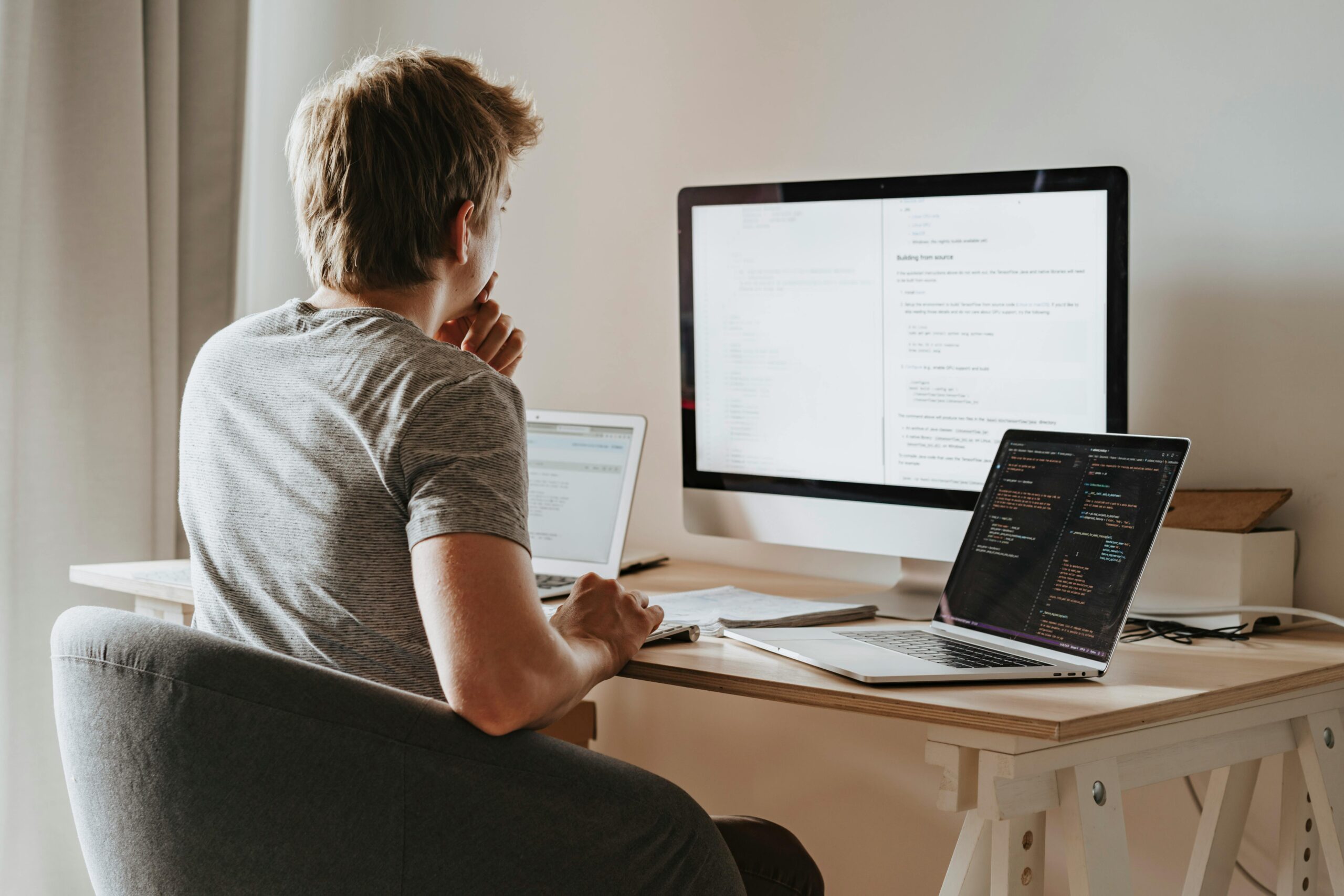 Eine Person sitzt an einem Schreibtisch mit einem Laptop und einem großen Desktop-Monitor, die beide Code oder technische Dokumente anzeigen. Ein weiterer offener Laptop in der Nähe zeigt ebenfalls Code an. Die Person wirkt konzentriert und nachdenklich.