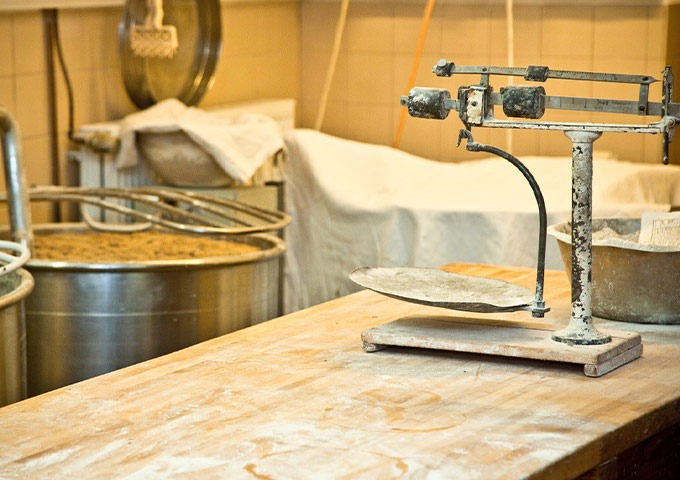 Eine rustikale Küchenszene mit einer alten Waage auf einem Holztisch, Mehl auf der Oberfläche und große Metallmischbehälter im Hintergrund.