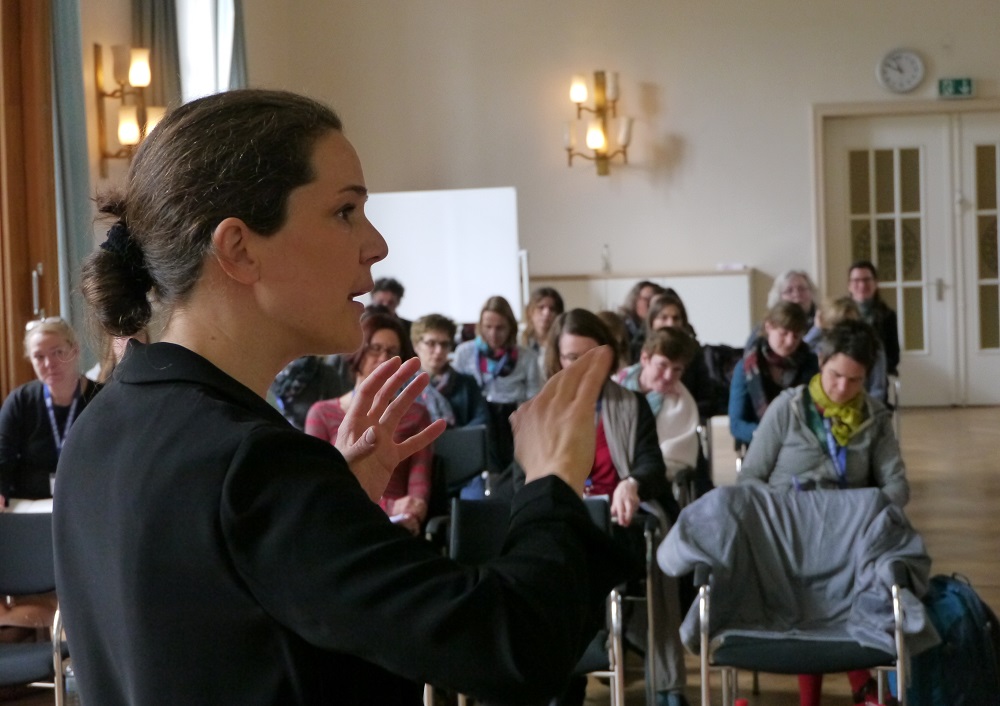 Eine Frau spricht vor einem sitzenden Publikum in einem hell erleuchteten Raum und gestikuliert mit ihren Händen, während die Zuhörer aufmerksam zuhören. Der Raum hat große Fenster, Wandleuchten und eine Uhr an der Wand.