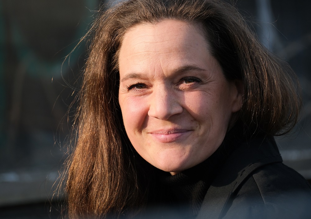 Eine Frau mit langen braunen Haaren und einem schwarzen Oberteil lächelt sanft in die Kamera bei weichem, natürlichem Licht, mit einem unscharfen Hintergrund im Freien.