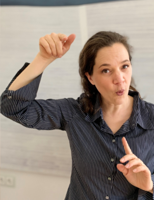 Eine Frau mit dunklem Haar, die ein gestreiftes Hemd trägt, gestikuliert ausdrucksstark mit einer erhobenen Hand, während die andere nach oben zeigt, als ob sie etwas dirigieren oder erklären würde. Sie scheint mitten im Satz zu sein.
