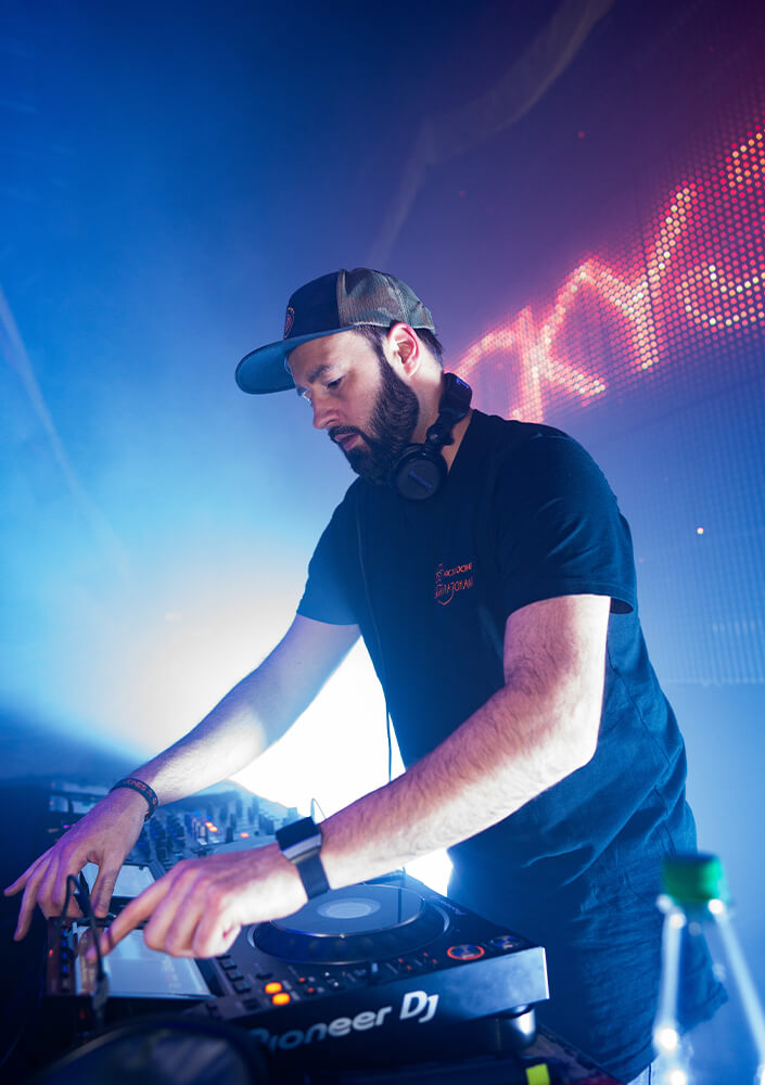 Ein DJ in schwarzem T-Shirt und Mütze tritt in einem Club auf, während er die Regler eines Pioneer-DJ-Mixers bedient, mit Kopfhörern um den Hals, bunten Bühnenlichtern und einem LED-Bildschirm im Hintergrund.