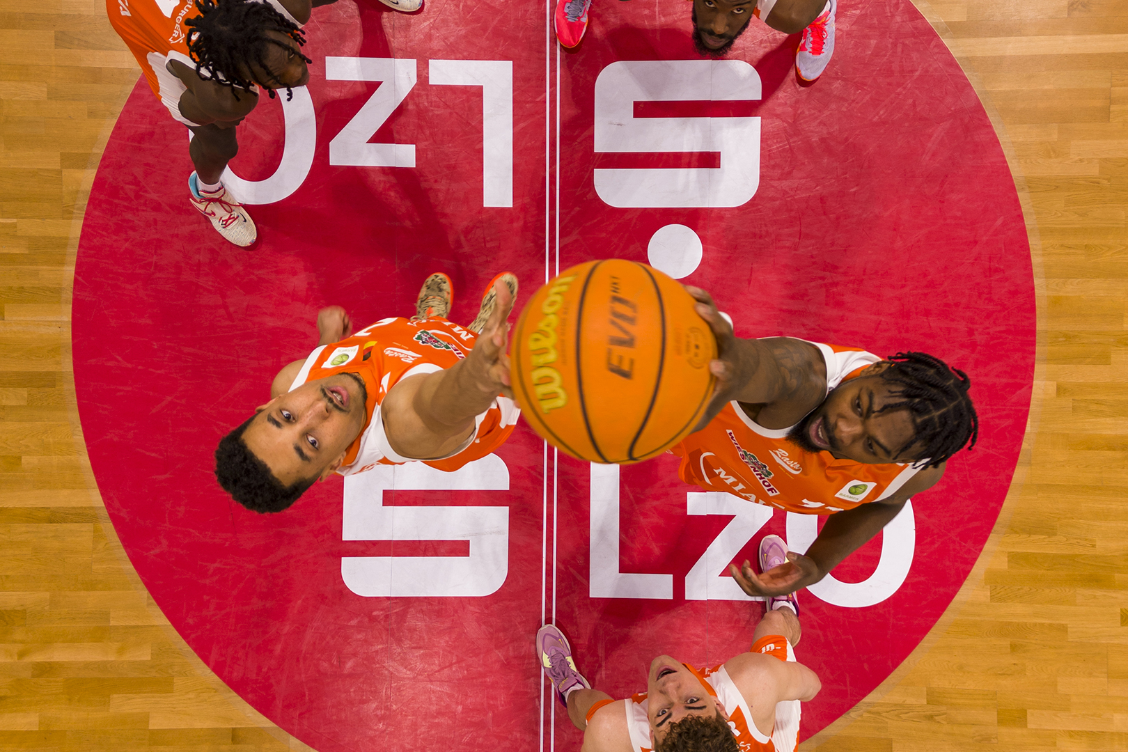 Draufsicht auf Basketballspieler auf einem Platz, die springen, um den Ball in der Mitte des Platzes zu erreichen, mit einem rot-weißen Kreis und einem Holzboden unter ihnen - so wird die Aufregung des Rasta Vechta Aufstiegs eingefangen.