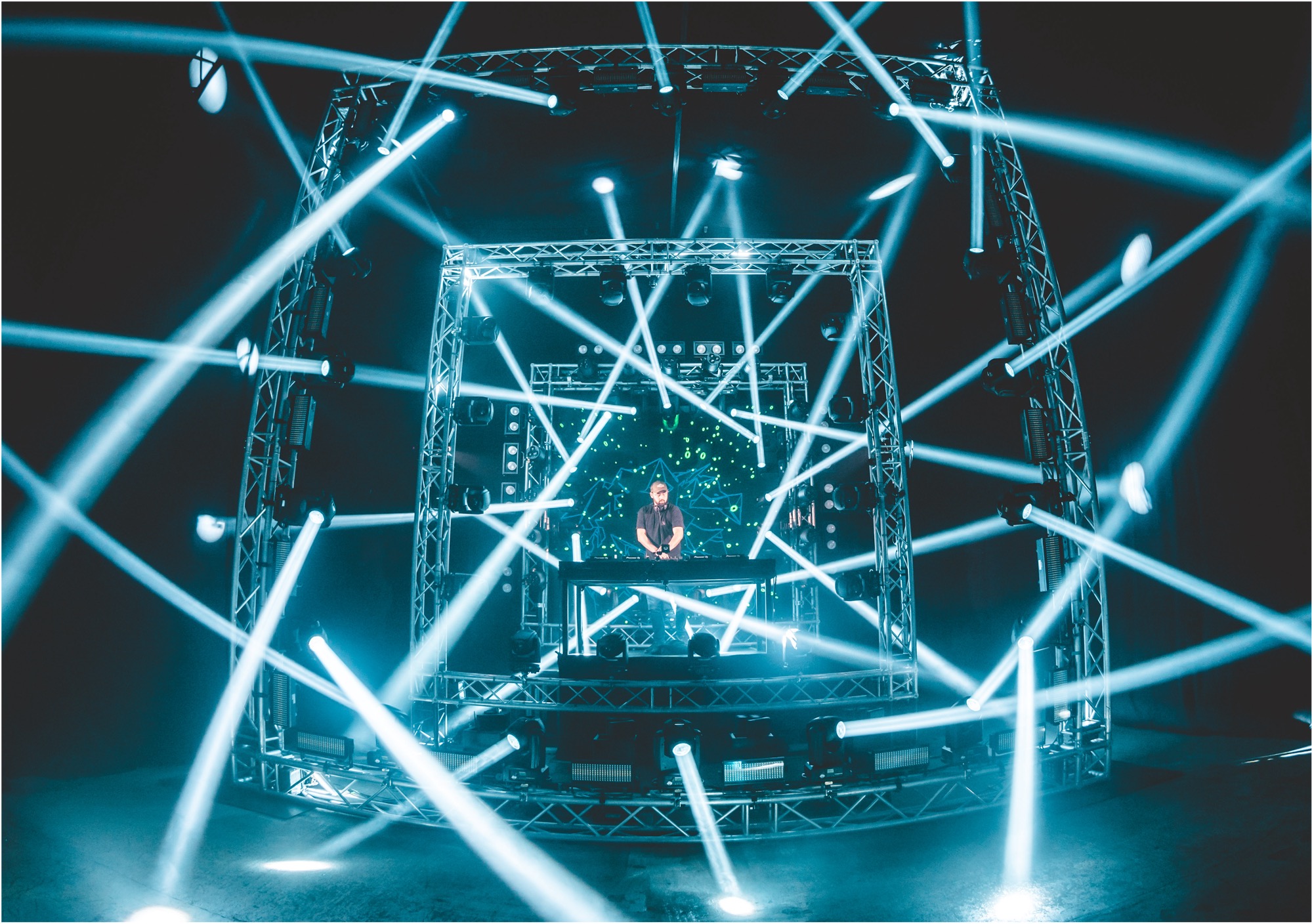 Ein DJ steht an einem Mischpult auf einer Bühne, die von einem Metallgerüst eingerahmt ist. Blaue Lichtstrahlen durchdringen den Rauch und schaffen eine dynamische, energiegeladene Atmosphäre bei der Oldenburger Land Nacht.