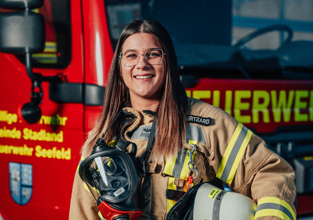 Eine lächelnde Feuerwehrfrau, Louisa Albertzard, mit Brille und voller Ausrüstung steht vor einem roten Fahrzeug der Freiwilligen Feuerwehr. Sie trägt einen Helm und ihr Namensschild lautet BERTZARD, passend zu den gelben und roten Buchstaben des Feuerwehrfahrzeugs.