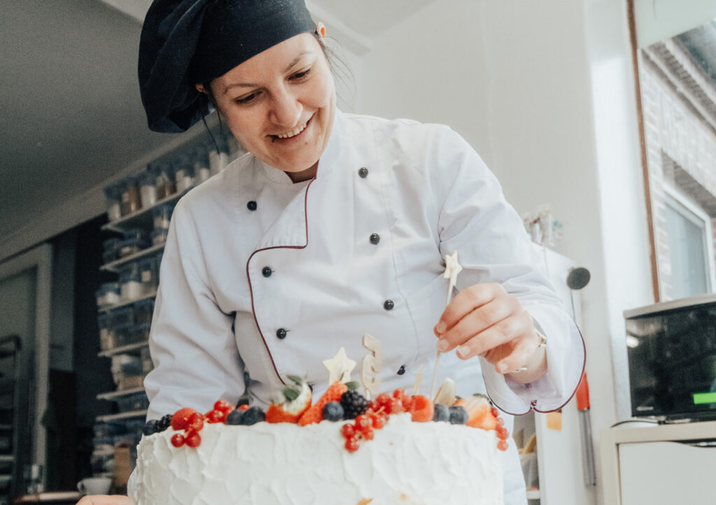 Ein lächelnder Konditor in weißer Uniform und schwarzer Mütze verziert in einer hellen Küche eine große, glasierte Torte mit Beeren und kleinen sternförmigen Aufsätzen.