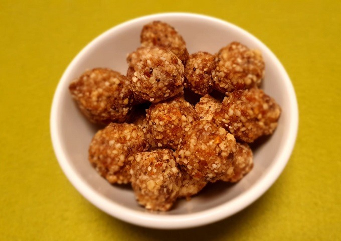Auf einer leuchtend gelben Fläche sitzt eine kleine weiße Schale, gefüllt mit runden, strukturierten Energiebällchen aus Nüssen und Samen - das perfekte Rezept für einen gesunden Snack.