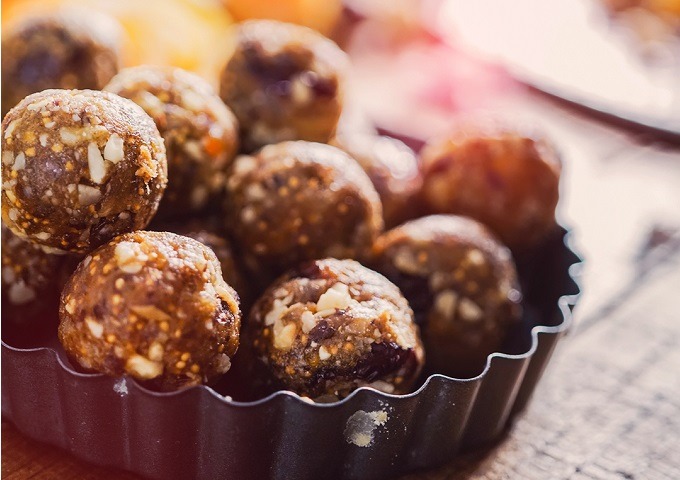 Nahaufnahme eines Tabletts, das mit runden, nussigen Energy Bites oder Snackballs gefüllt ist, auf denen Nüsse und Trockenfrüchte sichtbar sind. Die Oberfläche erscheint strukturiert und leicht glänzend, wobei das warme Licht die Details hervorhebt.