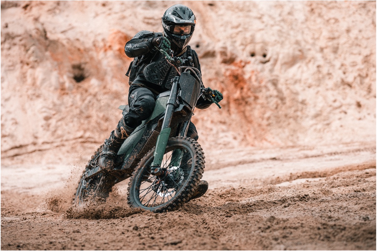 Eine Person, die schwarze Schutzkleidung und einen Helm trägt, fährt auf einem Geländemotorrad und wirbelt in einer scharfen Kurve auf einem zerklüfteten, sandigen Terrain Sand auf.