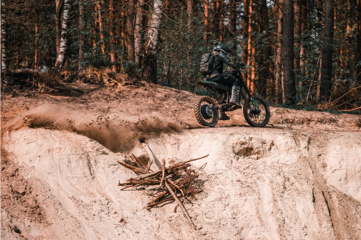 Eine Person, die einen Helm und einen Rucksack trägt, fährt mit einem Geländemotorrad schnell am Rande einer sandigen Klippe in einem Wald entlang und wirbelt dabei Staub in der Nähe eines Asthaufens auf.
