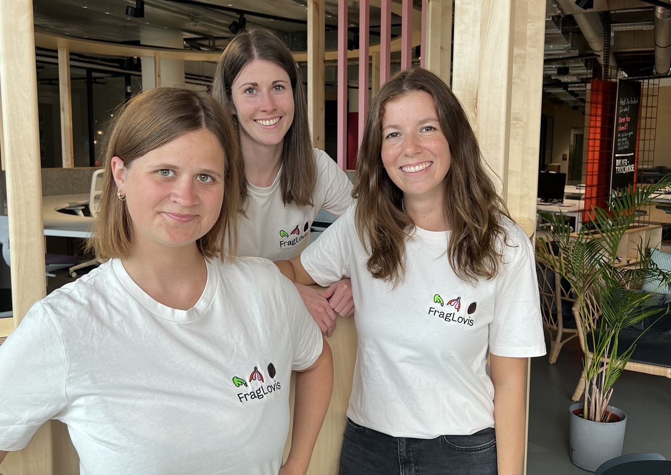 Drei lächelnde Frauen, die in einem Raum zusammenstehen und passende weiße T-Shirts mit dem FragLovis-Logo tragen. Der Hintergrund zeigt eine moderne Büroeinrichtung mit Holzelementen und Pflanzen.