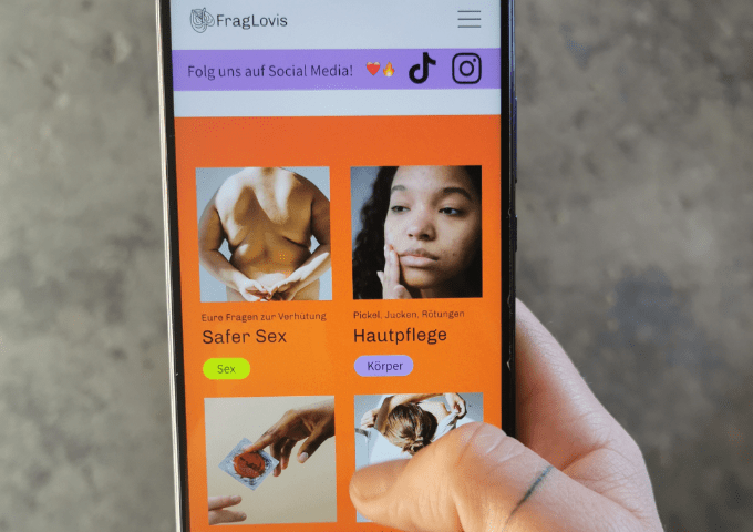 Eine Hand hält ein Telefon, das eine Website mit Themen zu sexueller Gesundheit und Hautpflege anzeigt, mit Bildern und deutschem Text zu "Safer Sex" und "Hautpflege". Oben sind Icons für soziale Medien zu sehen.