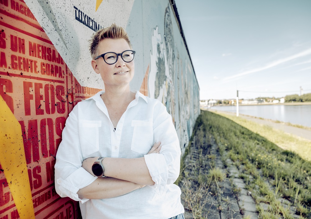 Eine Person mit kurzen Haaren und Brille, die ein weißes Hemd trägt, steht lächelnd mit verschränkten Armen vor einer bunten Graffiti-Wand in der Nähe eines Flussufers an einem sonnigen Tag.