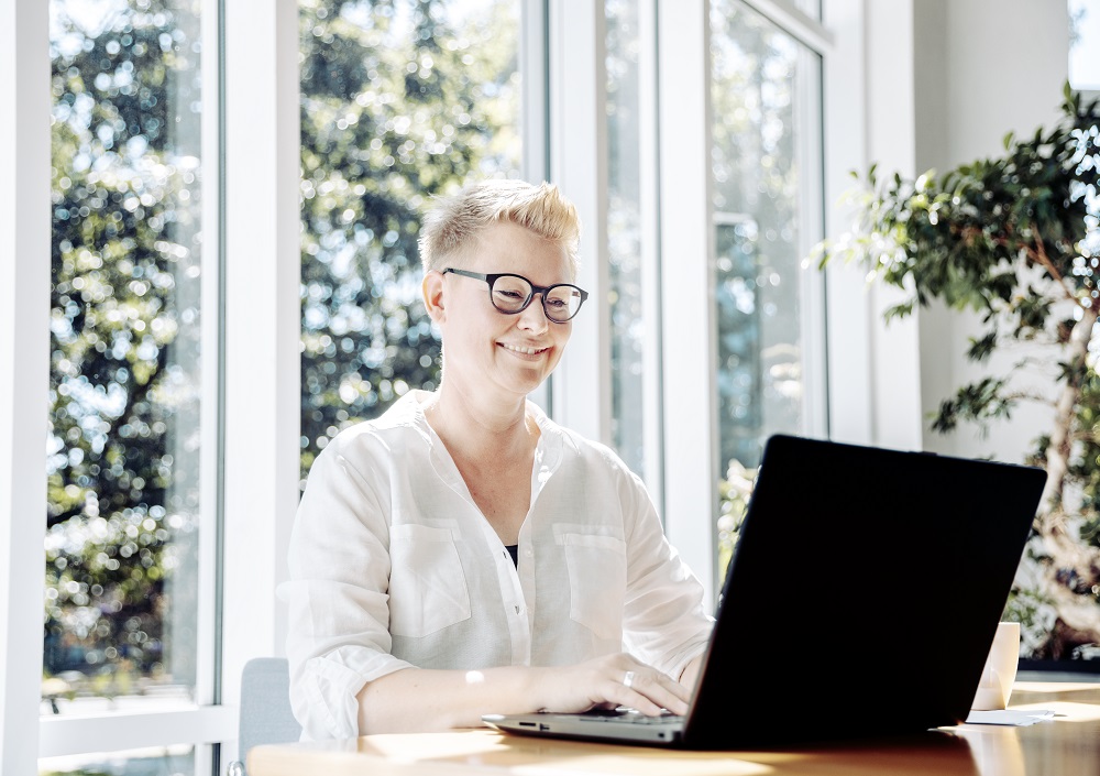 Eine lächelnde Person mit kurzen blonden Haaren und Brille arbeitet an einem Laptop an einem sonnenbeschienenen Tisch vor großen Fenstern, aus denen man Bäume sehen kann.