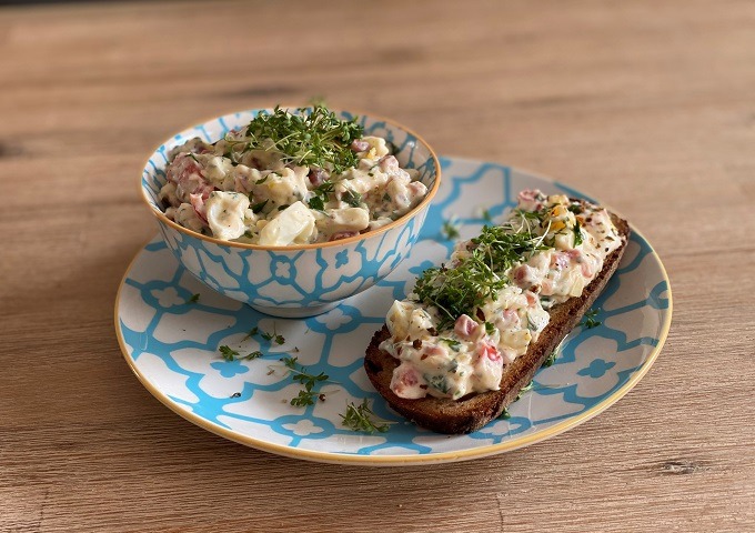 Eine Schale und eine Scheibe Brot, belegt mit einem cremigen Salat mit gewürfeltem Gemüse und Kräutern, serviert auf einem gemusterten Teller auf einer Holzunterlage.