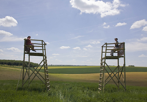 Zwei Personen sitzen sich auf hohen Holzplattformen in einem grasbewachsenen Feld gegenüber, unter einem blauen Himmel mit vereinzelten Wolken. Im Hintergrund erstrecken sich Ackerland und Bäume.