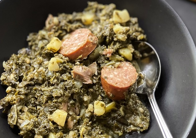 Nahaufnahme einer Schüssel mit einem deftigen Eintopf mit Blattgemüse, Kartoffelstückchen, Fleischstücken und Wurstscheiben, mit einem Löffel in der Schüssel.