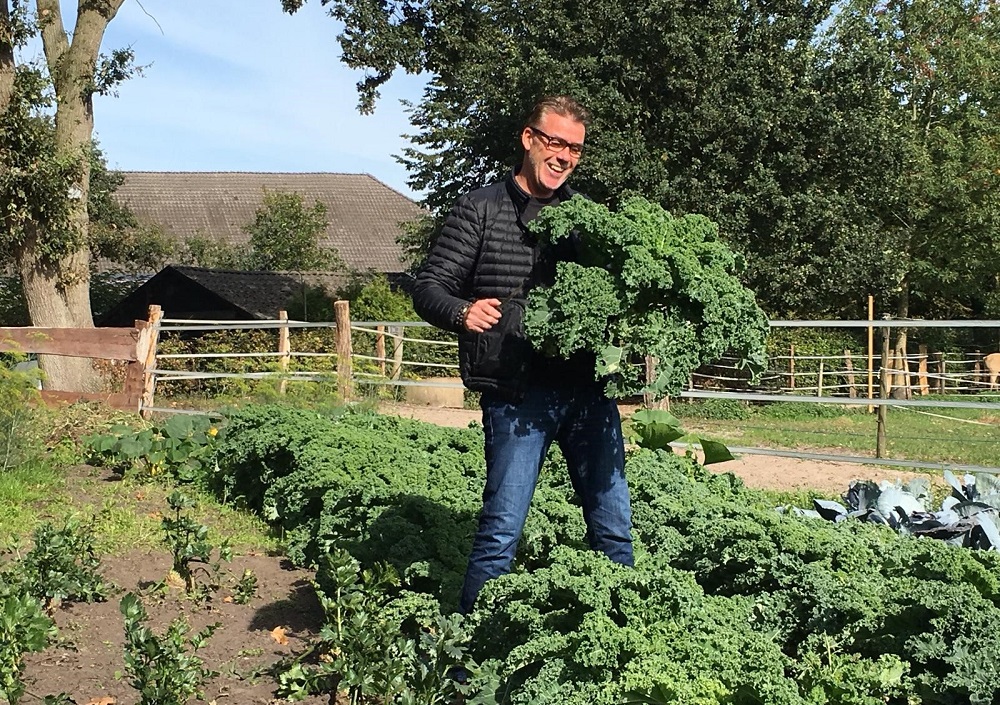 Ein Mann mit Brille und schwarzer Jacke steht in einem sonnigen Garten, lächelt und hält einen großen Strauß Grünkohl in der Hand, umgeben von Reihen von Grünkohlpflanzen und Bäumen im Hintergrund.