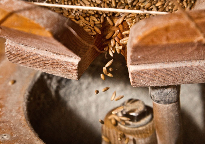 Nahaufnahme von Körnern, die aus einer hölzernen Rutsche in eine traditionelle Steinmühle fallen, um dort gemahlen zu werden.