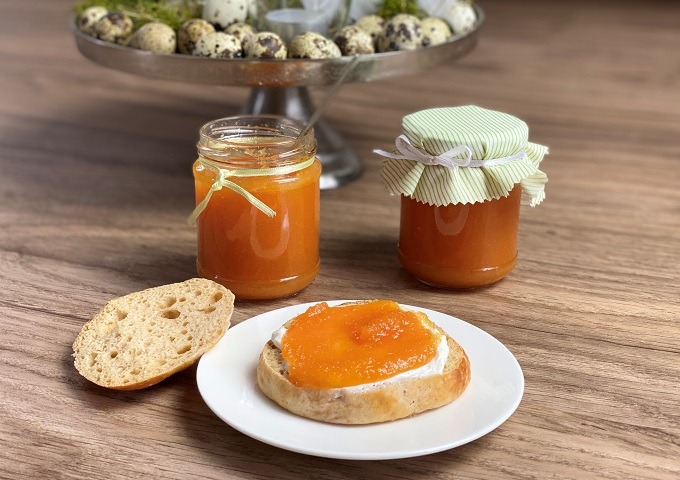 Ein Teller mit Brot, das mit Orangenmarmelade bestrichen ist, ein aufgeschnittenes Stück Brot, zwei Gläser mit Orangenmarmelade und ein Tablett mit Eiern im Hintergrund, alles auf einer hölzernen Fläche.