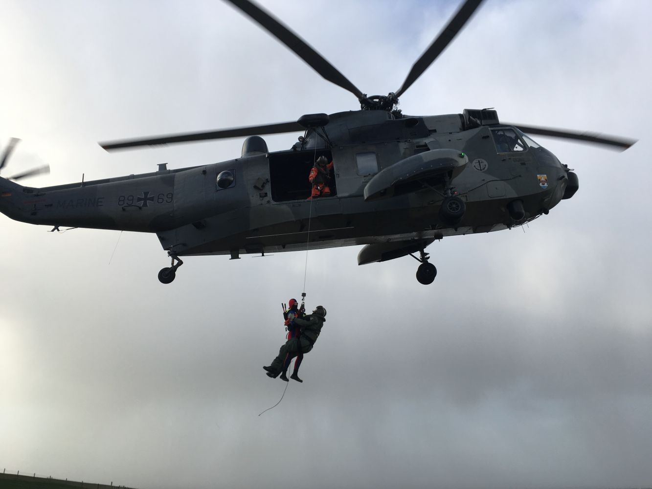 Ein Militärhubschrauber schwebt in der Luft, während eine Person an einem Rettungsseil hochgezogen wird, während eine andere Person von der Tür des Hubschraubers aus Hilfe leistet. Die Szene scheint eine Such- und Rettungsaktion darzustellen.